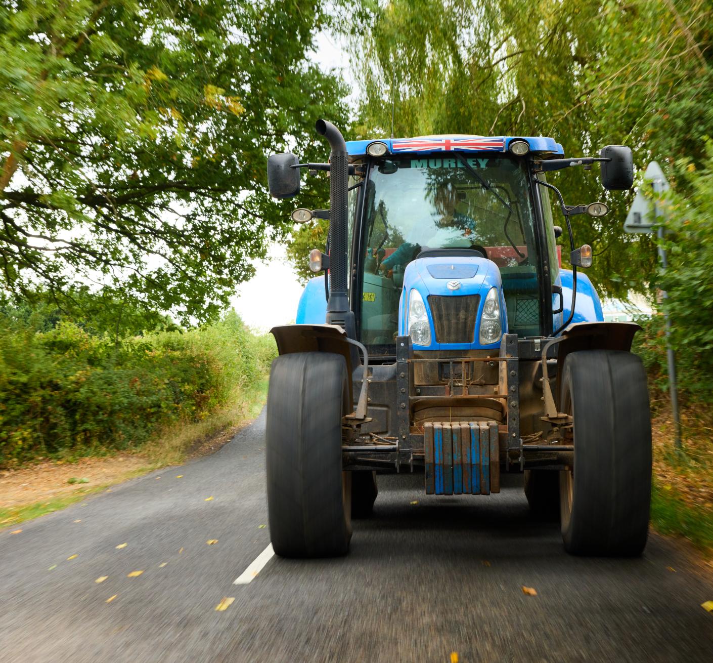 A blue tractor driving down a road lined with trees and bushes