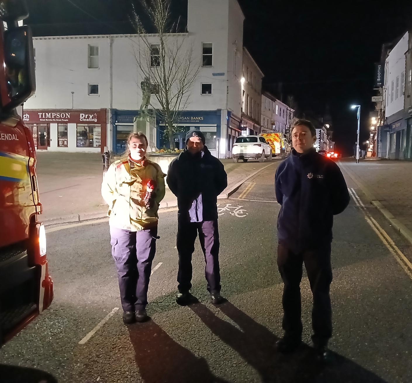 Three firefighters stand by a fire engine in the centre of a town