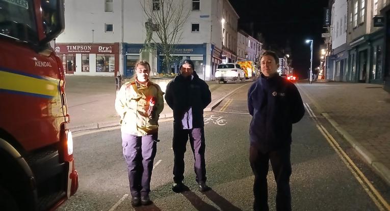 Three firefighters stand by a fire engine in the centre of a town