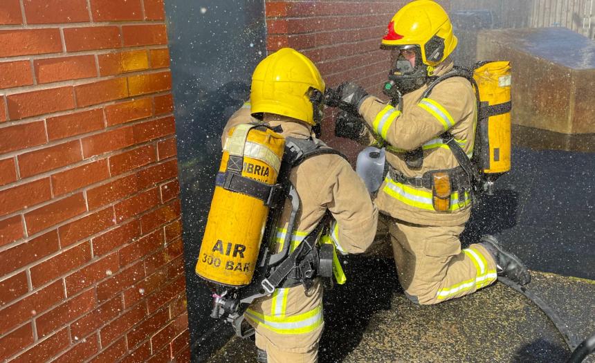 Two Firefighters in full PPE, completing a BA training session at a door, in a red brick building